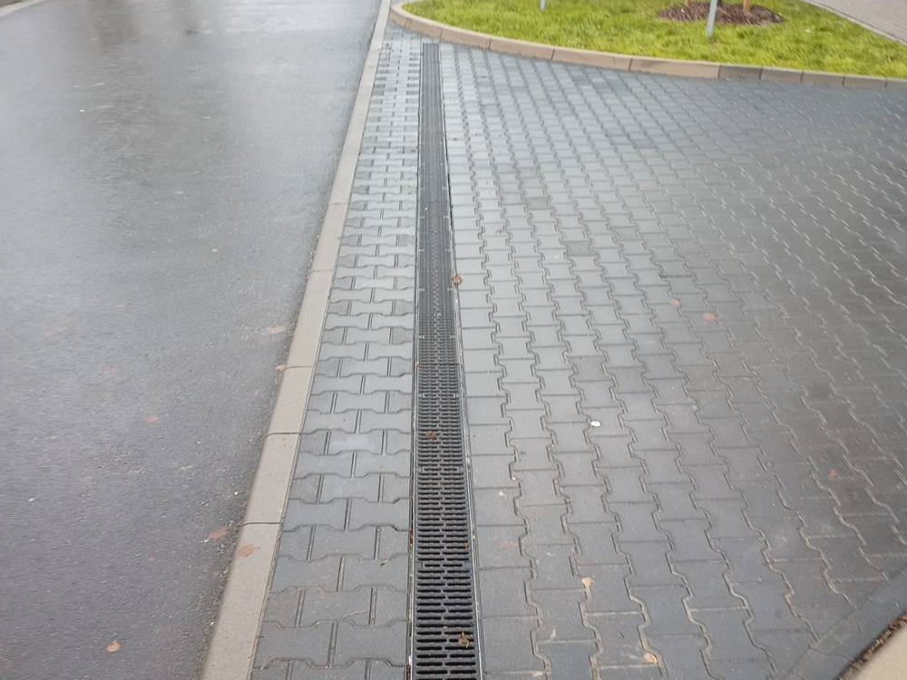 Odwodnienie liniowe z kratką obok chodnika z kostki brukowej i asfaltowej nawierzchni