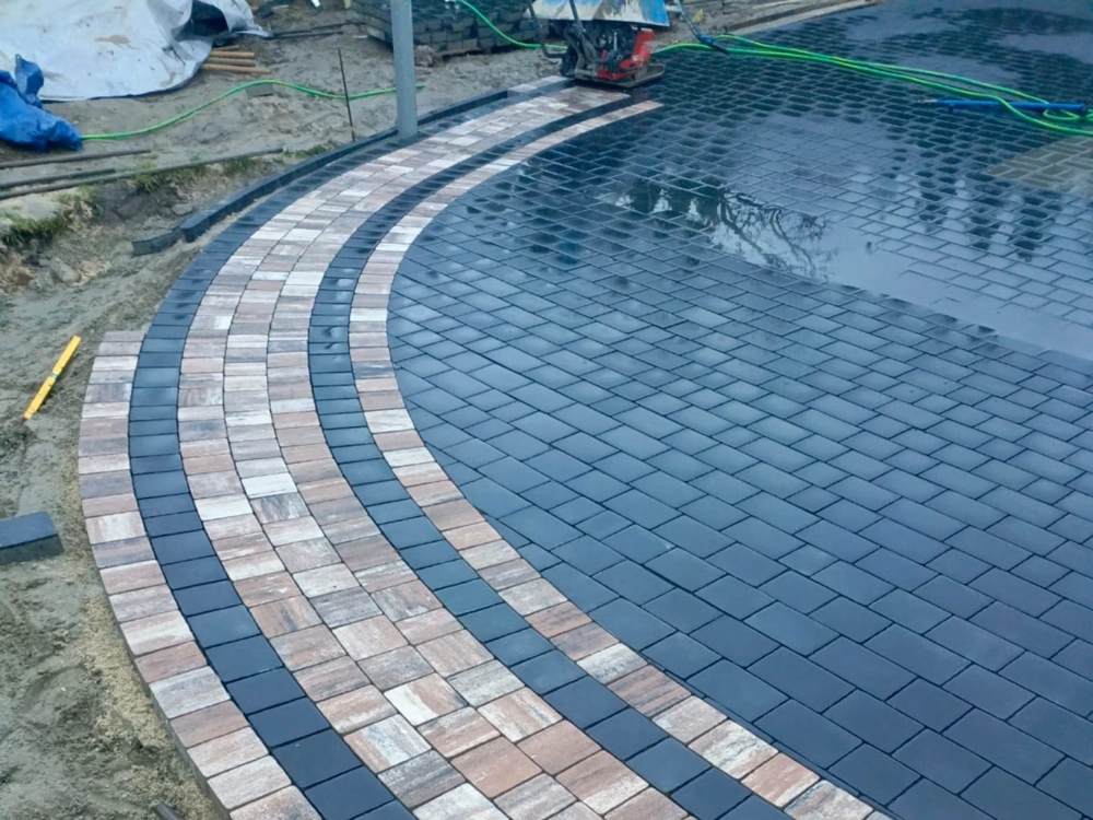 Układanie kostki brukowej – zwarta nawierzchnia z czarnych płytek i kontrastowych pasów z beżowo-brązowej kostki