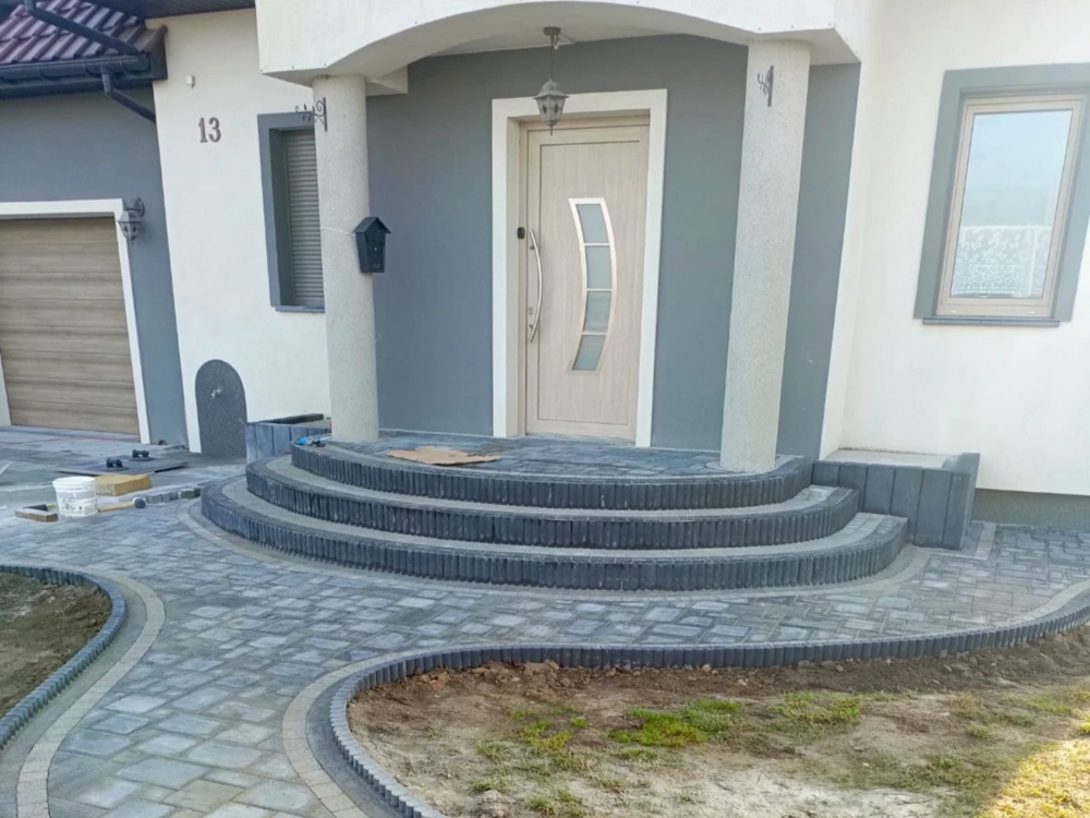 Schody z kostki brukowej frontowe – trzy półkoliste stopnie z szarej kostki i prowadzący chodnik z krawężnikami