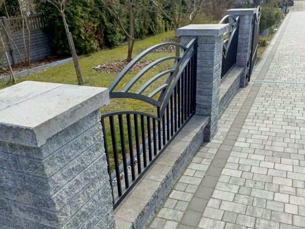 Ogrodzenie z bloczków z metalowymi przęsłami na podmurówce obok chodnika z kostki brukowej