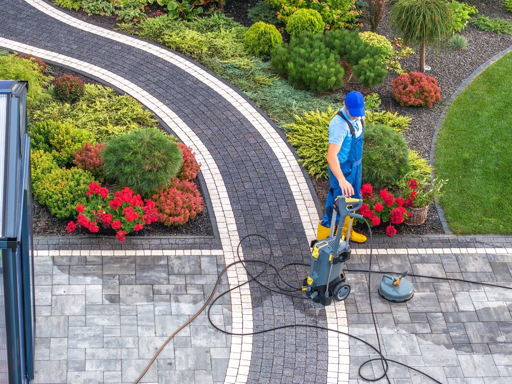 Pracownik czyści kostkę brukową myjką ciśnieniową na podjeździe otoczonym ozdobnymi rabatami