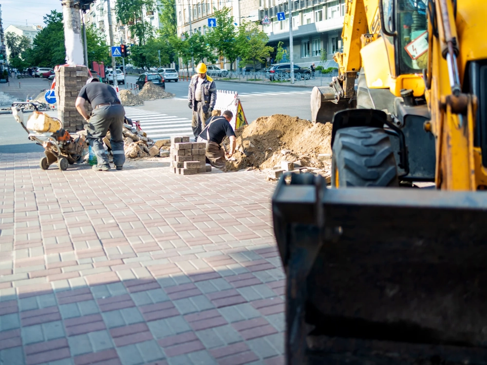 Układanie kostki brukowej na miejskim chodniku przez zespół brukarski z minikoparką i narzędziami