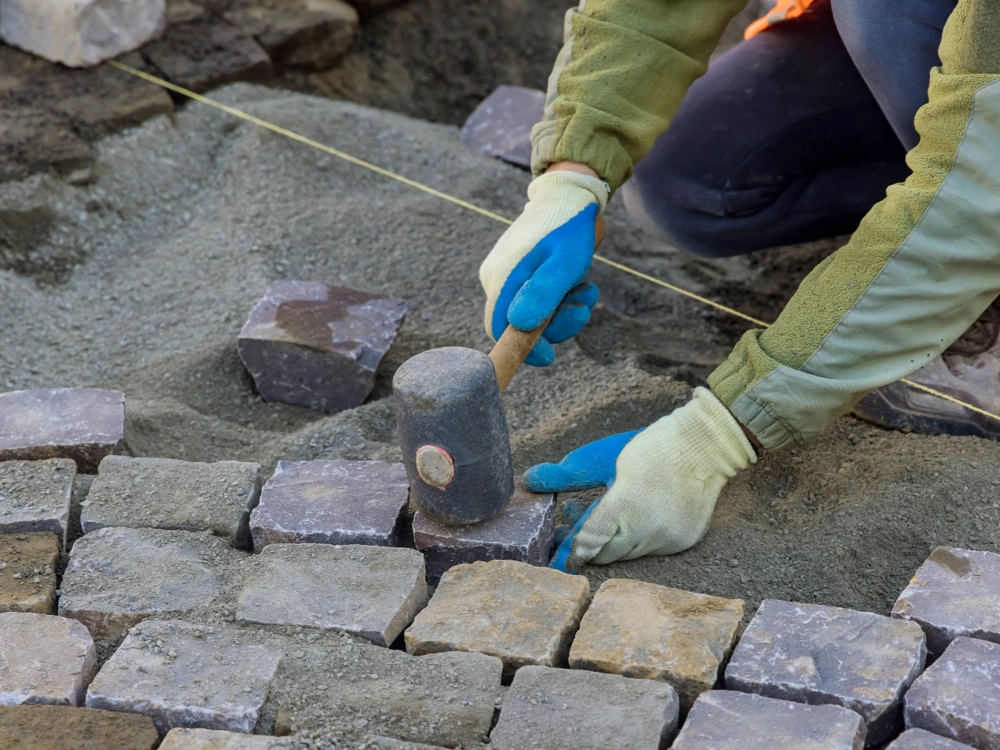 Układanie kostki brukowej, pracownik ustawiający kamienne granitowe płyty i wyrównujący je młotkiem na piaskowej podbudowie