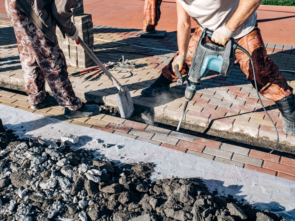 Demontaż starej nawierzchni i wymiana kostki brukowej młotem udarowym i łopatą przez robotników